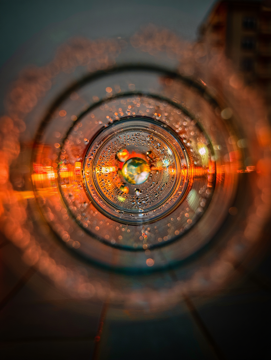 A macro photograph captures a unique perspective through water droplets, creating a tunnel-like view into a nighttime urban scene